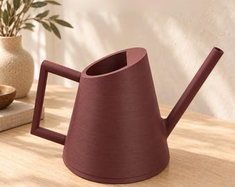 May include: A dark maroon watering can with a wide, tapered body, a long narrow spout, and a geometric angular handle. The surface features a subtle horizontal wood-grain texture. The text ATELIER LINEA HAUS is visible at the top.