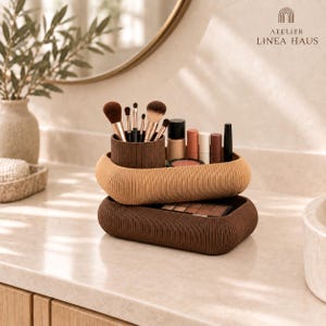 May include: Two stacked oval-shaped ribbed storage trays in light and dark brown tones. The top tray holds a cylindrical cup filled with various makeup brushes and several cosmetic tubes. The bottom tray contains a rectangular eyeshadow palette.