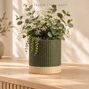 May include: A cylindrical dark green ceramic planter with a textured diamond pattern and a smooth light beige base. The planter holds a lush arrangement of eucalyptus branches, small white baby's breath flowers, and trailing green beads.