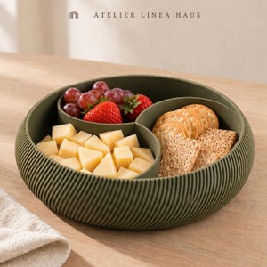 May include: A round, olive green serving tray with a ribbed exterior and three internal compartments. The tray contains red grapes, strawberries, cubed yellow cheese, round crackers, and rectangular whole-grain crackers. Text reads ATELIER LINEA HAUS.