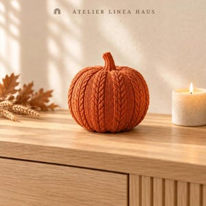 May include: A burnt orange decorative pumpkin featuring a textured cable knit pattern across its surface. The pumpkin has a prominent stem at the top and is positioned centrally. The text ATELIER LINEA HAUS is visible at the top of the image.