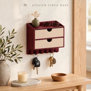 May include: A wall-mounted organizer by Atelier Linea Haus featuring a deep red frame with two beige drawers, a top shelf, and six hooks. The shelf holds a small vase with dried flowers and sunglasses. Keys hang from the hooks below.