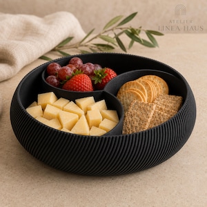 May include: A round black serving bowl with a ribbed exterior texture and three internal compartments. The bowl contains red grapes, fresh strawberries, cubed yellow cheese, round crackers, and square whole grain crackers arranged inside.