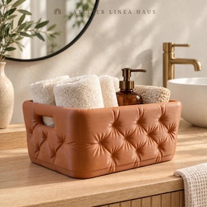 May include: A rectangular, terracotta-colored storage basket with a tufted, button-pressed exterior texture and integrated side handles. The basket contains several folded, cream-colored textured washcloths and a brown glass soap dispenser.