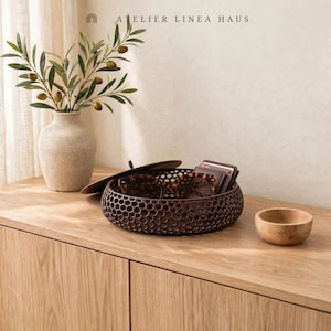 May include: A beige textured vase with olive branches, a dark brown circular honeycomb-patterned storage bowl with a lid, and a small wooden bowl. The text ATELIER LINEA HAUS is visible at the top. The items are arranged on a light wood surface.