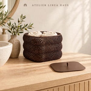 May include: A dark brown square storage basket with a textured honeycomb pattern, containing two rolled beige towels. Next to the basket is a matching flat square lid with a small central knob. The text ATELIER LINEA HAUS is visible.