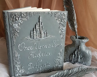 May include: A gray, textured book with a silver castle design and the text "Once Upon a Time Fredrick & Debbynie August 6th 2021". A silver feather quill pen and inkwell are also pictured.