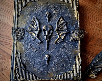 May include: A dark blue journal with a gold embossed cover featuring a skull and wings design. The journal has a distressed look with frayed edges and a rustic feel.