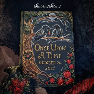 May include: A black book with a gold inscription that reads "Once Upon A Time" and "October 20, 2023". The book is decorated with a tree, two black birds, and red roses.