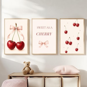 May include: A set of three wall art prints featuring a light cream background. The left print shows two red cherries tied with a pink ribbon. The center print displays the text SWEET AS A CHERRY above a pink ribbon. The right print shows scattered cherries.