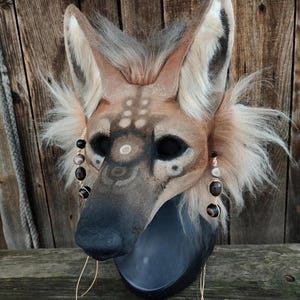 Puede incluir: Una máscara de hiena detallada y hecha a mano con un diseño realista. La máscara presenta una combinación de colores marrón y beige con detalles en negro alrededor de los ojos y la nariz. Tiene orejas grandes y esponjosas y está adornada con pendientes de cuentas y detalles de alambre dorado. La máscara está montada sobre una base negra.