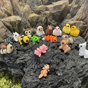 Op de afbeelding: Een verzameling miniatuur dierenfiguren, waaronder een grijze konijn, een witte lam, een bruine uil, een gele bij, een groene kikker en een roze varken. Andere dieren zijn een bruine koe, een witte kip en gele eendjes. De beeldjes staan op een donkere rots.