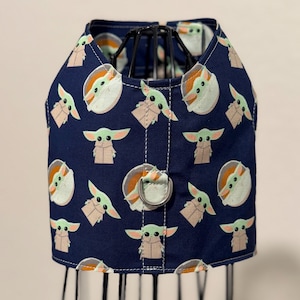 May include: A navy blue dog harness featuring a repeating pattern of a cartoon character with large ears and a tan robe. The harness has a metal D-ring for leash attachment and white stitching.