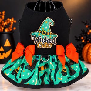 May include: A black pet harness dress featuring a teal witch hat graphic with the text Wicked Cute, two bright orange bows at the waist, and a teal skirt patterned with black witch boots, crescent moons, and white stars on a black trim.