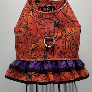 May include: A Halloween-themed dog harness featuring a vibrant orange and red spiderweb pattern. The harness includes a purple ruffled skirt with black trim and a silver metal ring for leash attachment. The design incorporates black spider accents.