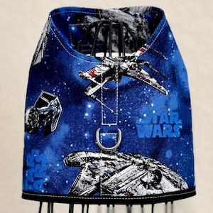 May include: A blue dog harness featuring a space-themed design with illustrations of spaceships and the text "STAR WARS". The harness has a D-ring for leash attachment and a black trim.