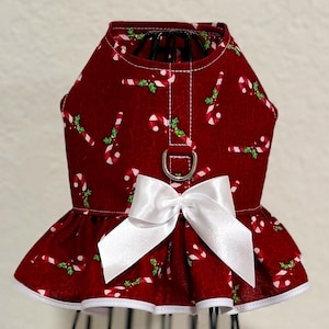 May include: A red dog dress with a white bow and candy cane pattern. The dress has a ruffled skirt and a D-ring for leash attachment. The trim is white.