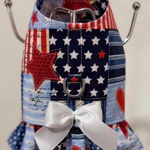 May include: A patriotic dog harness featuring a patchwork design with red, white, and blue fabrics. The harness includes a large red star, white stars on a navy background, and a white bow. It has a metal D-ring for leash attachment.