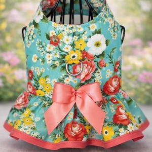 May include: A floral dog harness with a turquoise background and a pattern of red, yellow, and white flowers. The harness has a pink bow and trim. It features a metal ring for leash attachment.
