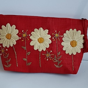 Peut inclure: Une trousse de maquillage rouge avec des fleurs de marguerite brodées. Les marguerites ont des pétales crème et des centres jaunes. Le sac a une tirette à pompon et est fait d'un tissu texturé.