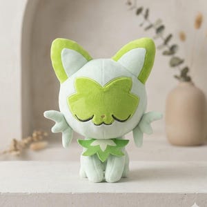May include: A plush toy cat in a seated position, featuring a light green and white color scheme. The cat has closed eyes, a green face mask, and green accents on its ears and collar. The toy has small, wing-like appendages on its sides.