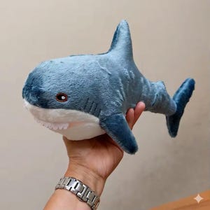 May include: A plush shark toy featuring a soft blue upper body and a white underbelly. The stuffed animal has embroidered brown eyes, visible white teeth along the mouth, a dorsal fin, and a tail fin, crafted from a fuzzy fabric material.