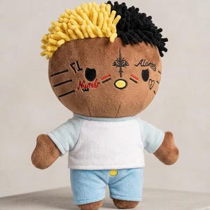 May include: A plush doll with a brown face, yellow and black hair, and embroidered facial details. The doll wears a white and light blue shirt and blue shorts. The face has the word "Numb" and the word "Alone" embroidered on it.