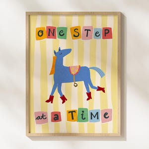 May include: A framed art print featuring a blue horse wearing red boots and a saddle, set against a yellow and white striped background. The text "one step at a time" is displayed in colorful blocks.
