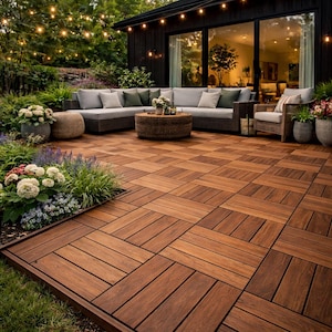 Może przedstawiać: Zewnętrzne patio z ciemną drewnianą podłogą z płytek. Szara sofa narożna z poduszkami i okrągły wiklinowy stolik kawowy. Czarny budynek z dużymi szklanymi drzwiami w tle, z wiszącymi lampkami.