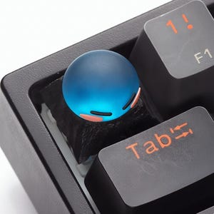 Peut inclure: Un capuchon de touche sphérique bleu avec un motif de visage de dessin animé, posé sur un clavier mécanique noir. Le capuchon de touche a un effet dégradé et de petits accents roses. Les touches du clavier sont noires avec des lettres orange.