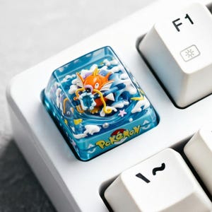 May include: A custom keycap featuring a 3D design of a cartoon orange and yellow creature with blue and white accents, set against a blue background with the word "Pokémon". The keycap is on a white keyboard.