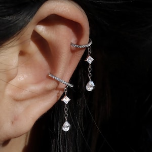 May include: Silver ear cuffs with dangling cubic zirconia gemstones. One cuff curves around the upper ear, while the other is a straight bar. Each cuff has a chain with a square and teardrop-shaped gemstone.