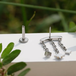 Peut inclure: Une paire de boucles d'oreilles argent&eacute;es. Une boucle d'oreille est compos&eacute;e d'une tige avec un disque plat, l'autre d'une barre incurv&eacute;e avec trois cha&icirc;nes pendantes. Chaque cha&icirc;ne se termine par un petit charme en forme d'&eacute;toile.