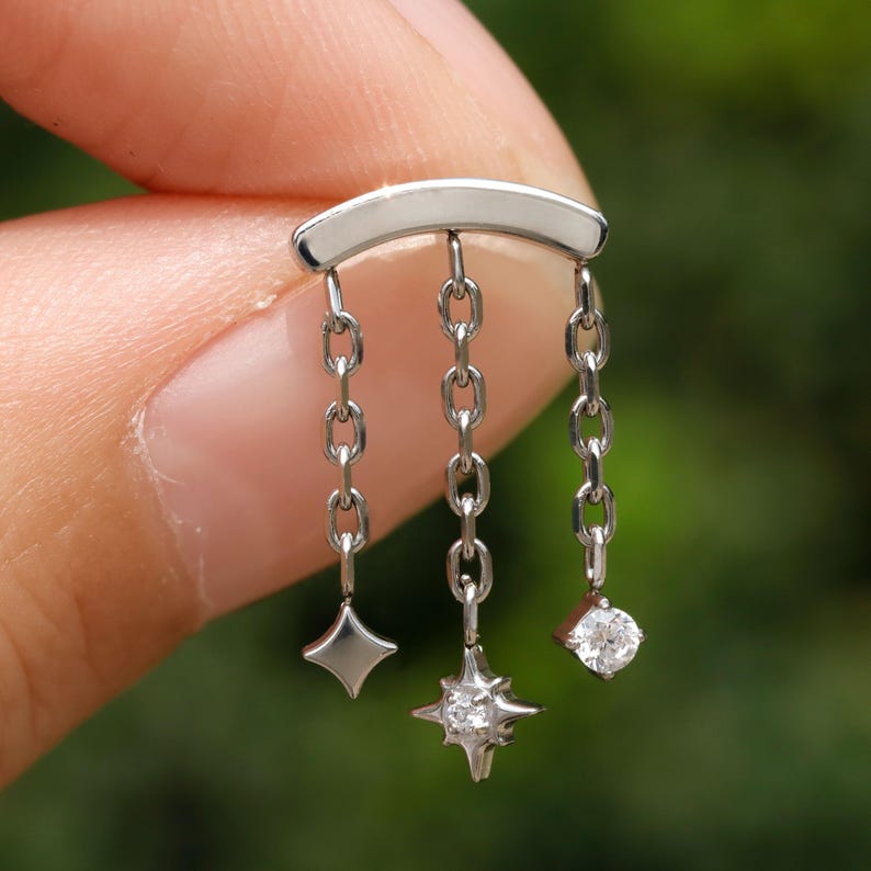 Peut inclure: Boucle d'oreille argent&eacute;e avec une barre sup&eacute;rieure incurv&eacute;e et trois cha&icirc;nes pendantes. Chaque cha&icirc;ne porte un charme diff&eacute;rent : un losange, une &eacute;toile avec une gemme centrale et une gemme ronde. La boucle est tenue entre les doigts sur un fond vert flou.