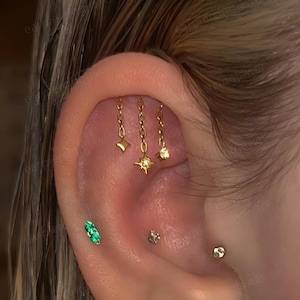 May include: A gold ear cartilage piercing featuring three dangling chains with star-shaped charms. Below, a double green gemstone stud, a small silver-toned stud, and a single gold-toned ball stud are positioned along the lower ear cartilage.
