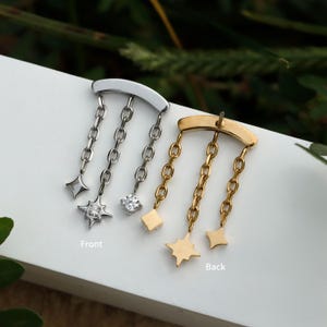 Peut inclure: Deux boucles d'oreilles, une argent&eacute;e et une dor&eacute;e, chacune avec une barre incurv&eacute;e et des cha&icirc;nes pendantes. La boucle d'oreille argent&eacute;e a un charm en forme de losange, un charm en forme d'&eacute;toile et une gemme transparente. La boucle d'oreille dor&eacute;e a un charm carr&eacute; et deux charms en forme d'&eacute;toile.