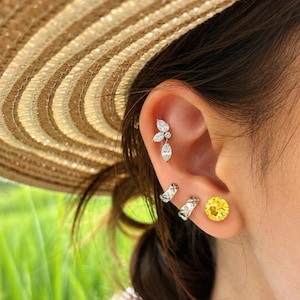 Può includere: Un orecchino da cartilagine color argento con tre pietre chiare taglio marquise disposte a grappolo a forma di foglia, accompagnato da due piccoli cerchi color argento con motivo testurizzato e un orecchino rotondo giallo brillante.