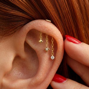 Puede incluir: Primer plano de un piercing de oreja dorado con tres colgantes colgantes: una estrella, una estrella fugaz con una gema central y una pequeña gema redonda. La joya está sujeta a una cadena. La oreja tiene un pequeño aro dorado en la parte superior.