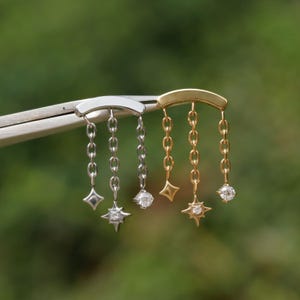 Peut inclure: Gros plan de deux paires de boucles d'oreilles. Une paire est argent&eacute;e, l'autre dor&eacute;e. Chaque boucle d'oreille a une barre incurv&eacute;e avec des cha&icirc;nes pendantes. Les cha&icirc;nes se terminent par des breloques en forme d'&eacute;toile et de diamant.