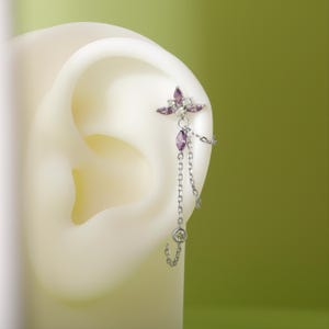 Puede incluir: Un piercing de oreja plateado con un diseño floral con piedras preciosas moradas y transparentes. El pendiente tiene una cadena con una gema morada colgante y una gema transparente al final. Se muestra sobre un modelo de oreja blanca.