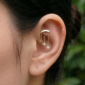 May include: A gold ear cartilage piercing jewelry piece featuring a curved bar with three dangling chains of varying lengths. The chains are adorned with small star-shaped charms and tiny clear round gemstones, creating a delicate layered look.