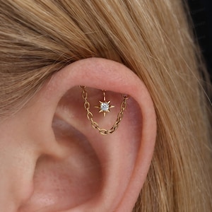 Peut inclure: Un piercing de cartilage d'oreille doré composé d'une fine chaîne reliant deux points, avec une breloque centrale en forme d'étoile sertie d'une pierre transparente. Le bijou est placé dans le pli supérieur du cartilage de l'oreille.