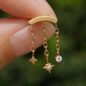 Peut inclure: Boucle d'oreille dor&eacute;e avec une barre incurv&eacute;e. Trois cha&icirc;nes pendent de la barre, chacune avec un charme : un losange, une &eacute;toile avec une petite gemme et une gemme ronde.