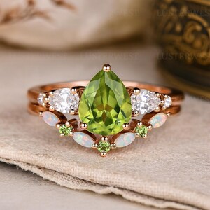 May include: A rose gold ring set featuring a pear-shaped, light green peridot center stone. The ring is accented with white gemstones and smaller green stones, with a delicate, layered design.