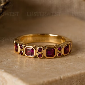 May include: A gold ring featuring a repeating pattern of square and round red gemstones. The ring is set on a neutral-toned surface, with a blurred background. The ring's design is symmetrical, with alternating shapes of gemstones.