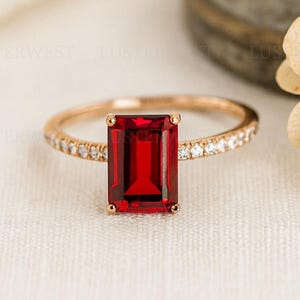 May include: A rose gold ring featuring a rectangular, deep red gemstone as the focal point. The band is adorned with small, sparkling accent stones. The ring is set against a soft, neutral background, highlighting its elegant design.