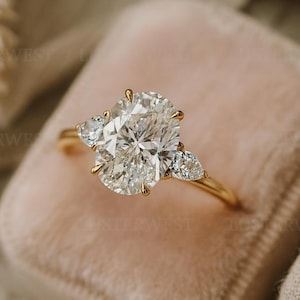May include: A gold engagement ring featuring an oval-cut diamond flanked by two pear-shaped diamonds. The ring is displayed on a pink velvet ring box.