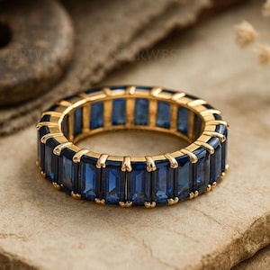 Peut inclure: Une bague dorée ornée d'une rangée de pierres précieuses rectangulaires bleu foncé. La bague est posée sur une surface texturée brun clair, avec un arrière-plan flou. Le design de la bague présente une rangée continue de pierres bleues.