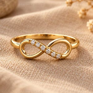 May include: A gold ring featuring an infinity symbol design. One side of the infinity loop is set with five small, clear, round-cut stones. The band is polished gold, creating a continuous, smooth, and reflective surface throughout the entire piece.