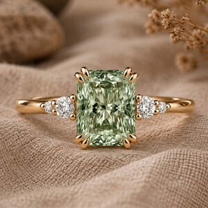Op de afbeelding: Een gouden ring met een grote, rechthoekige, lichtgroene middensteen die door vier klauwen wordt vastgehouden. De band is aan weerszijden versierd met twee kleinere, ronde, heldere stenen, wat zorgt voor een symmetrisch ontwerp met een gepolijste afwerking.